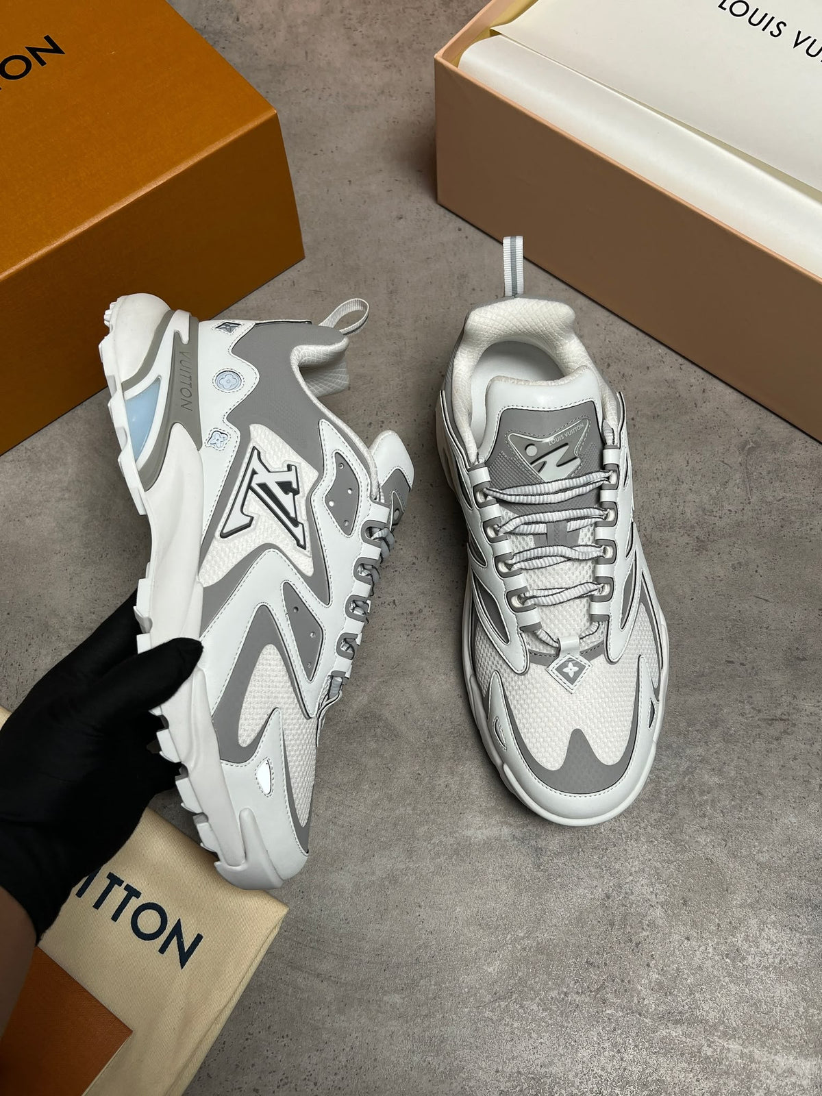 A pair of white and gray Louis Vuitton chunky sneakers, with one held by a black-gloved hand and the other placed on a gray floor. The orange and beige Louis Vuitton boxes and dust bag are visible in the background.