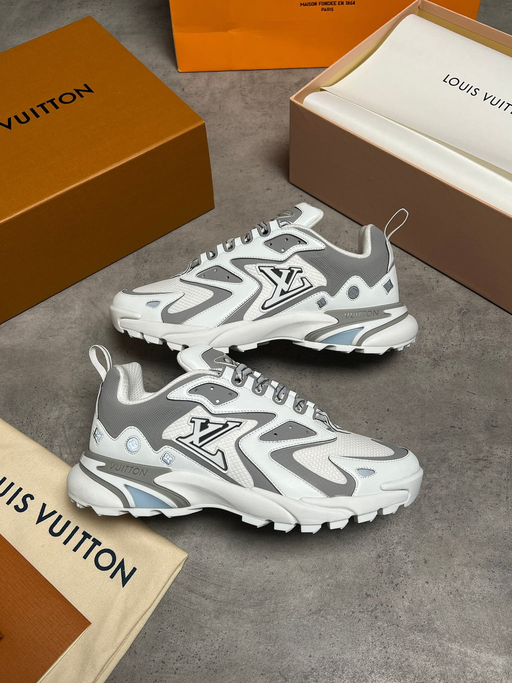 A pair of Louis Vuitton chunky sneakers in white and gray, displayed on a gray concrete surface with Louis Vuitton boxes and dust bags around them.