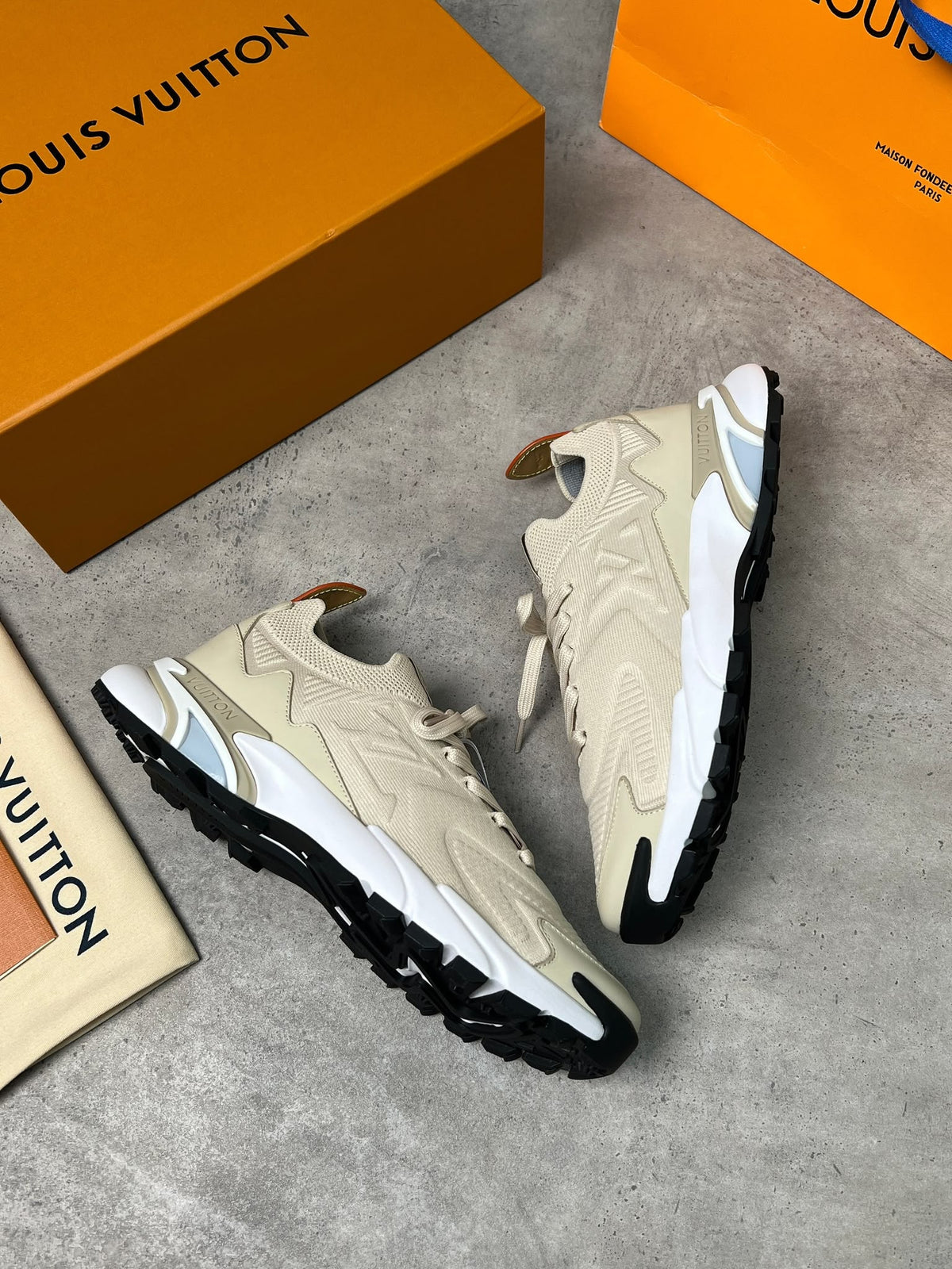 A pair of Louis Vuitton low-top sneakers in beige, white, and black, displayed on a gray concrete surface alongside orange Louis Vuitton boxes and a dust bag.