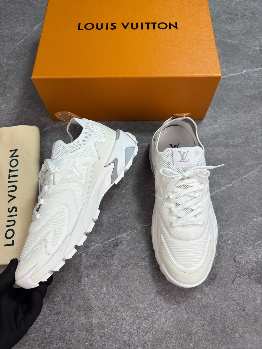 A pair of white Louis Vuitton sneakers displayed next to their signature orange box, dust bag, and a hand holding one shoe, all set on a gray stone floor.