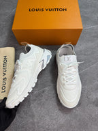 A pair of white Louis Vuitton sneakers displayed next to their signature orange box, dust bag, and a hand holding one shoe, all set on a gray stone floor.