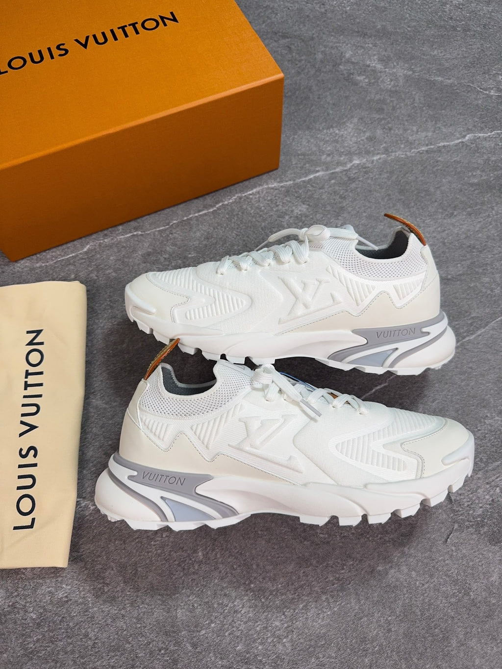 A pair of white Louis Vuitton low-top sneakers displayed on a gray stone-textured surface, with the orange Louis Vuitton shoebox and a beige dust bag visible in the background.