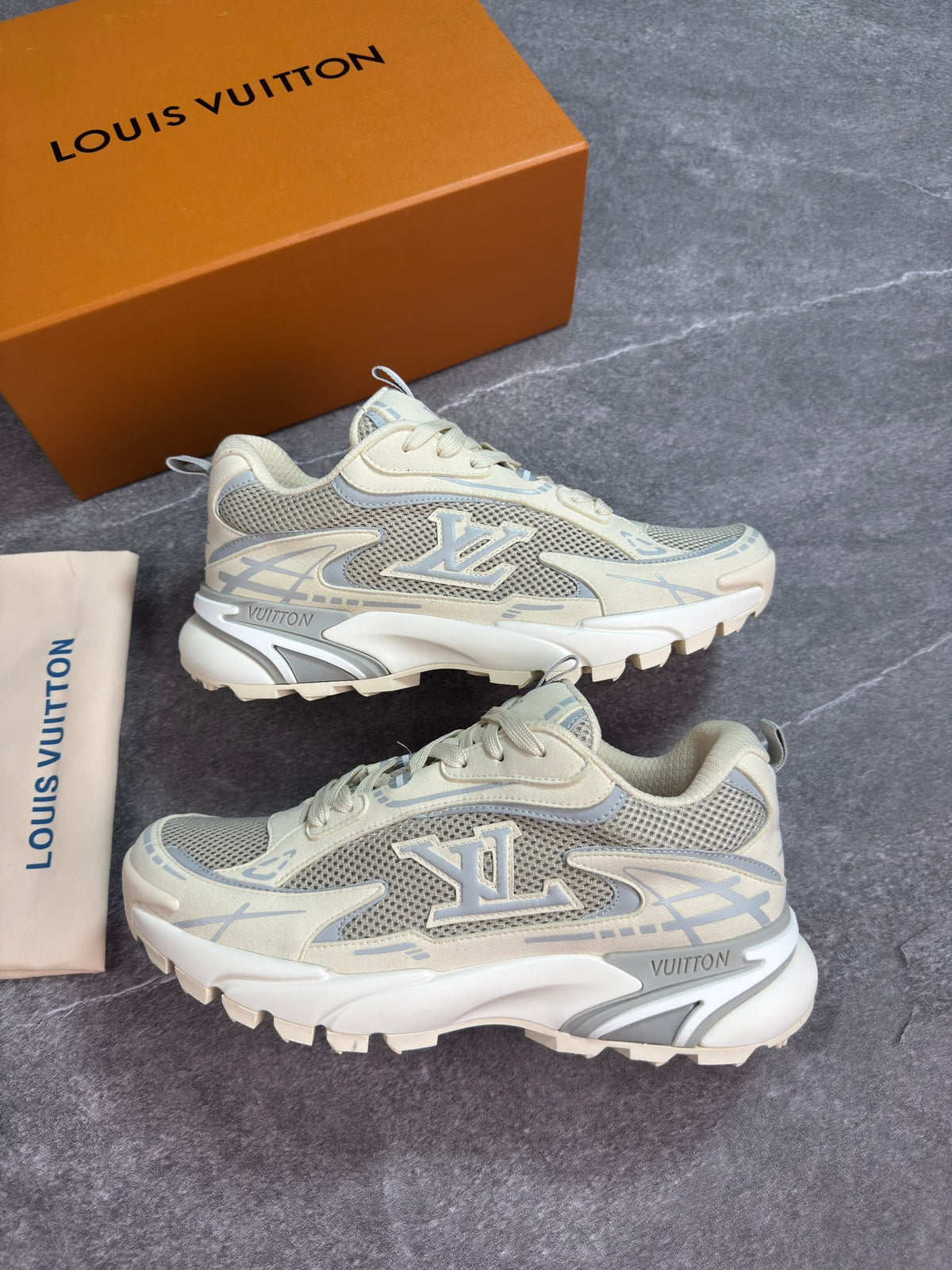 A pair of Louis Vuitton chunky sneakers in beige and silver-gray mesh, displayed next to their signature orange Louis Vuitton box and a beige dust bag on a gray stone-textured background.