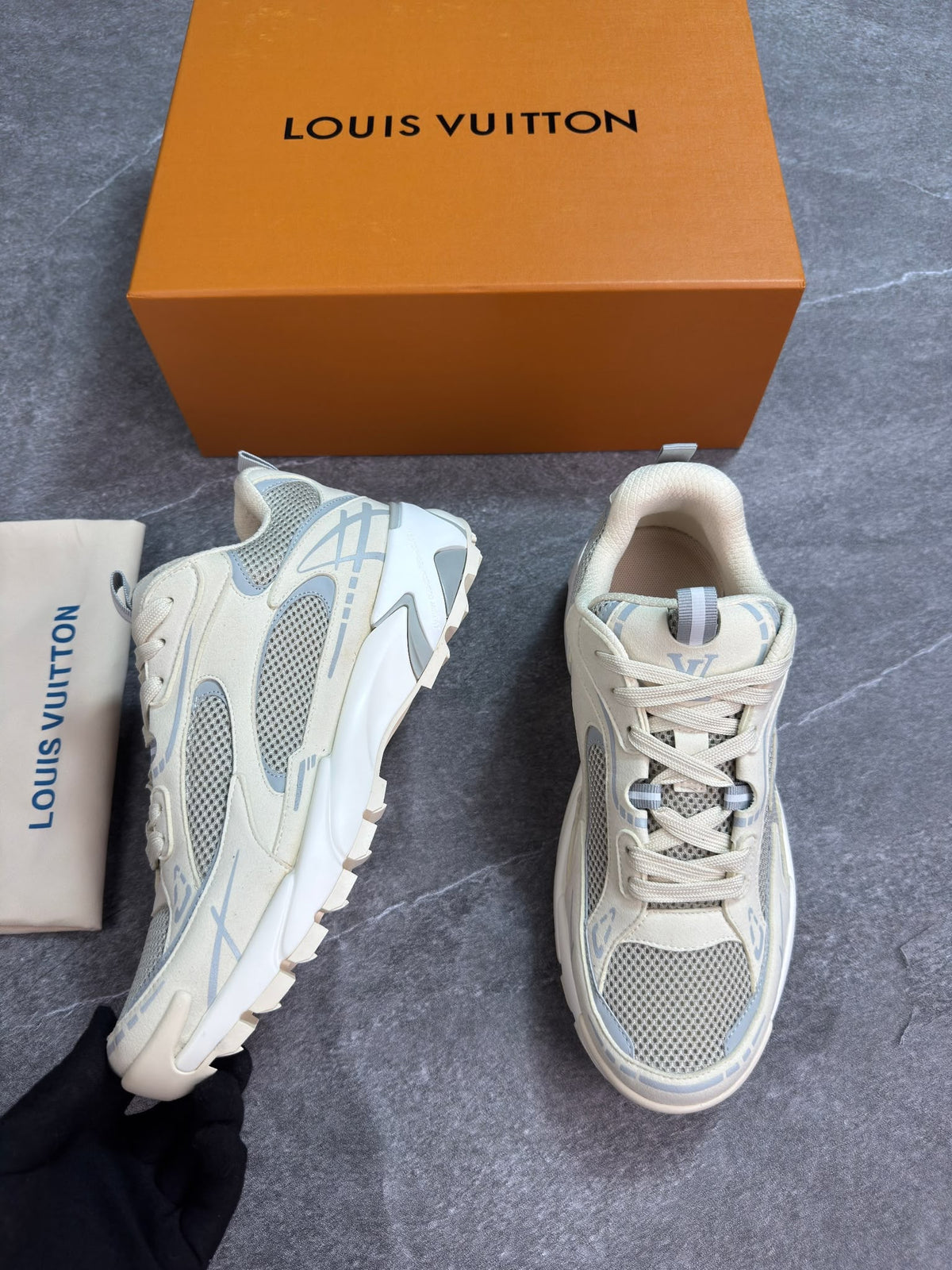 A pair of Louis Vuitton chunky sneakers in off-white and light gray, displayed next to their signature orange box and dust bag on a gray textured floor.