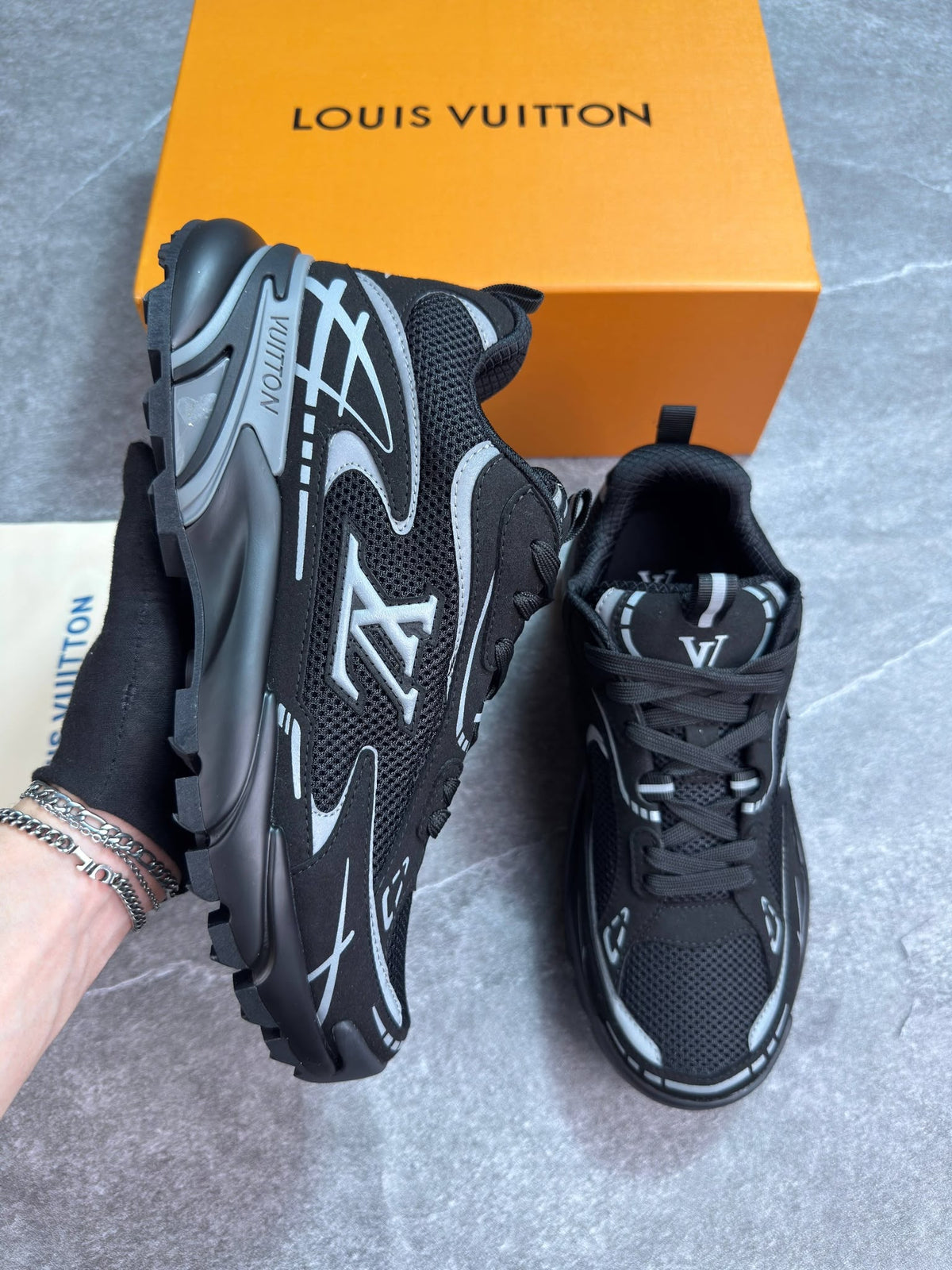 A pair of black and gray Louis Vuitton chunky sneakers shown with an orange Louis Vuitton box on a gray tiled floor. One shoe is held up by a hand to display the side profile, while the other sits on the floor.