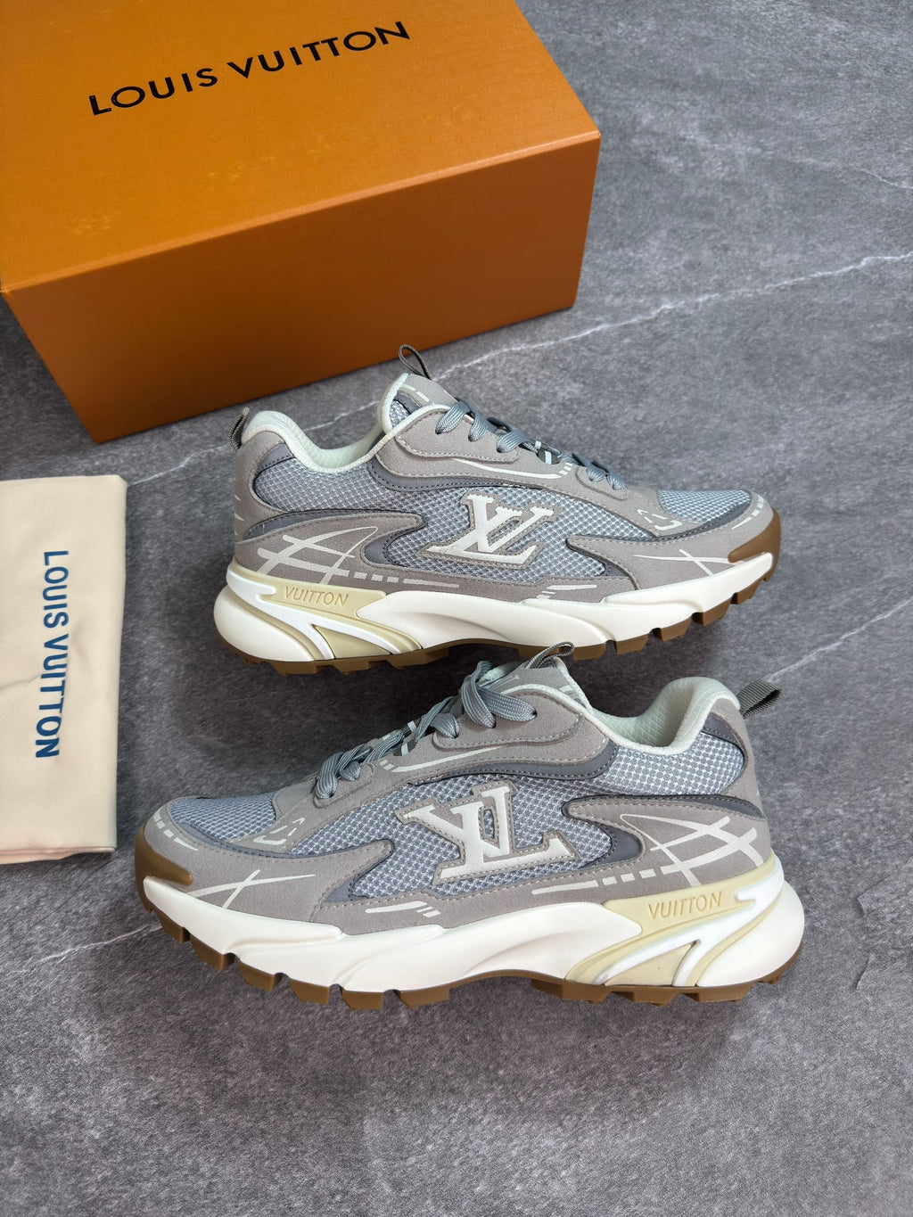 A pair of Louis Vuitton low-top sneakers in gray and white mesh with gold accents, displayed next to their orange brand box and dust bag on a gray stone surface.