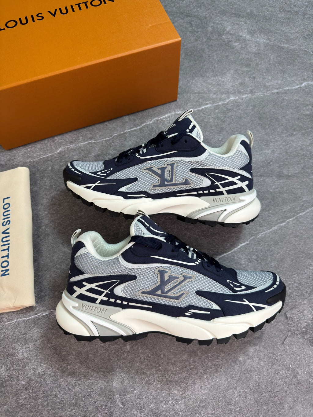 A pair of Louis Vuitton Air Max 2013 sneakers in navy blue, gray, and white, displayed next to their signature orange box and dust bag on a gray stone-textured background.