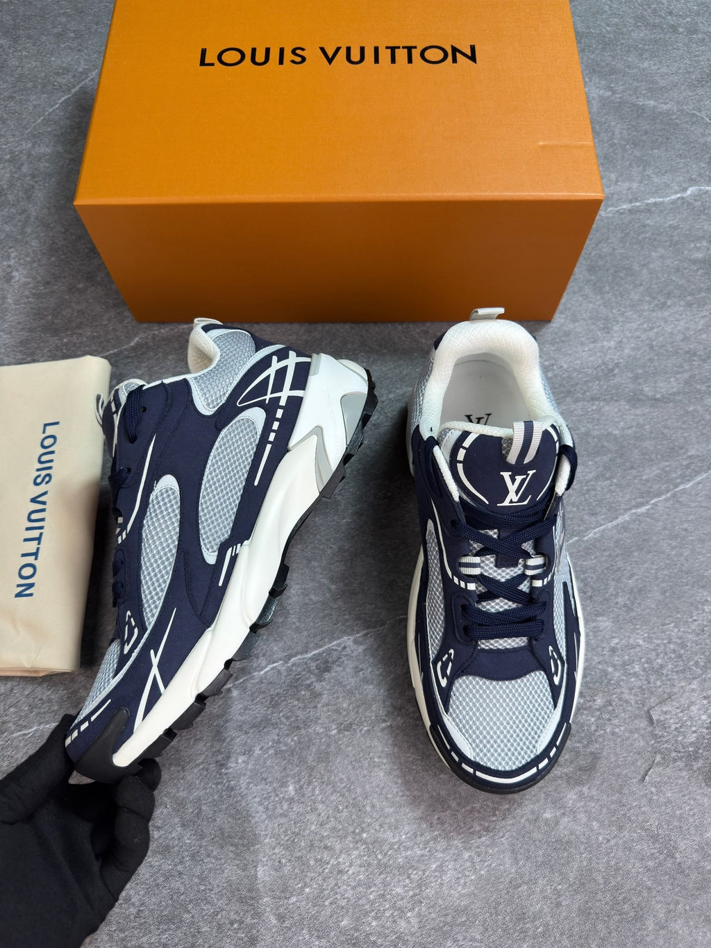 A pair of Louis Vuitton chunky sneakers in navy blue, white, and gray mesh, shown with an orange Louis Vuitton box, a dust bag, and a black gloved hand on a gray stone floor.