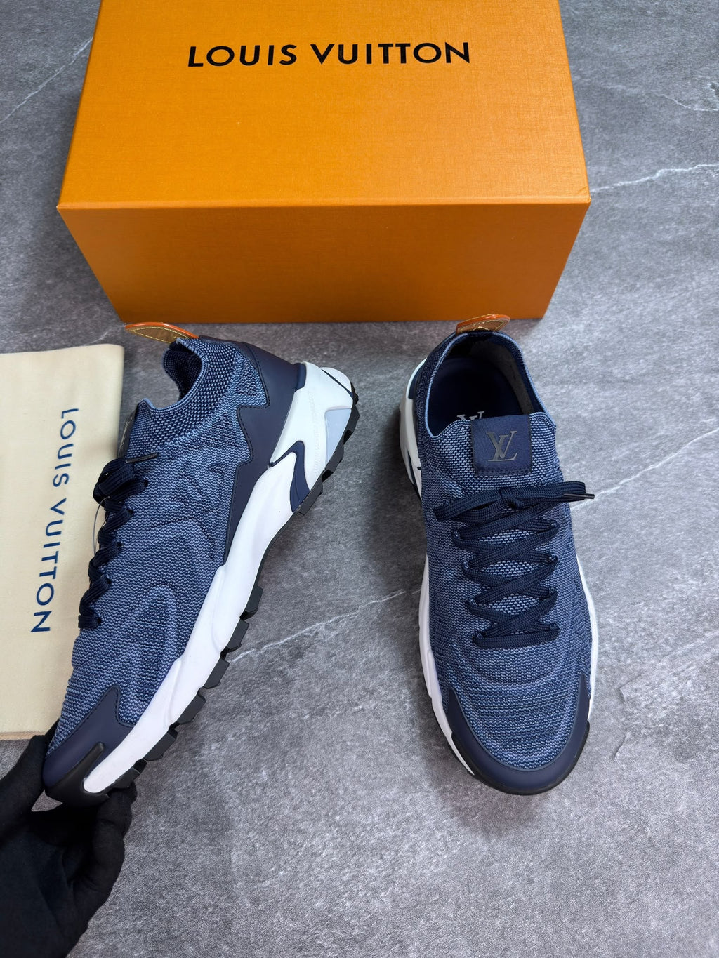 A pair of Louis Vuitton sneakers in navy blue and white, displayed next to their signature orange Louis Vuitton box and a beige dust bag on a gray stone floor.