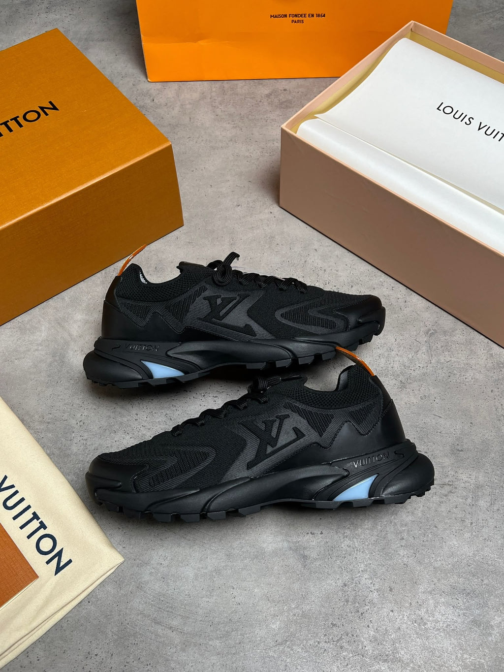 A pair of black Louis Vuitton sneakers displayed on a gray concrete surface, surrounded by their signature orange and beige Louis Vuitton packaging boxes and dust bags.