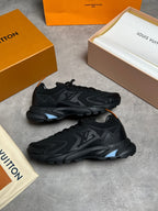A pair of black Louis Vuitton sneakers displayed on a gray concrete surface, surrounded by their signature orange and beige Louis Vuitton packaging boxes and dust bags.
