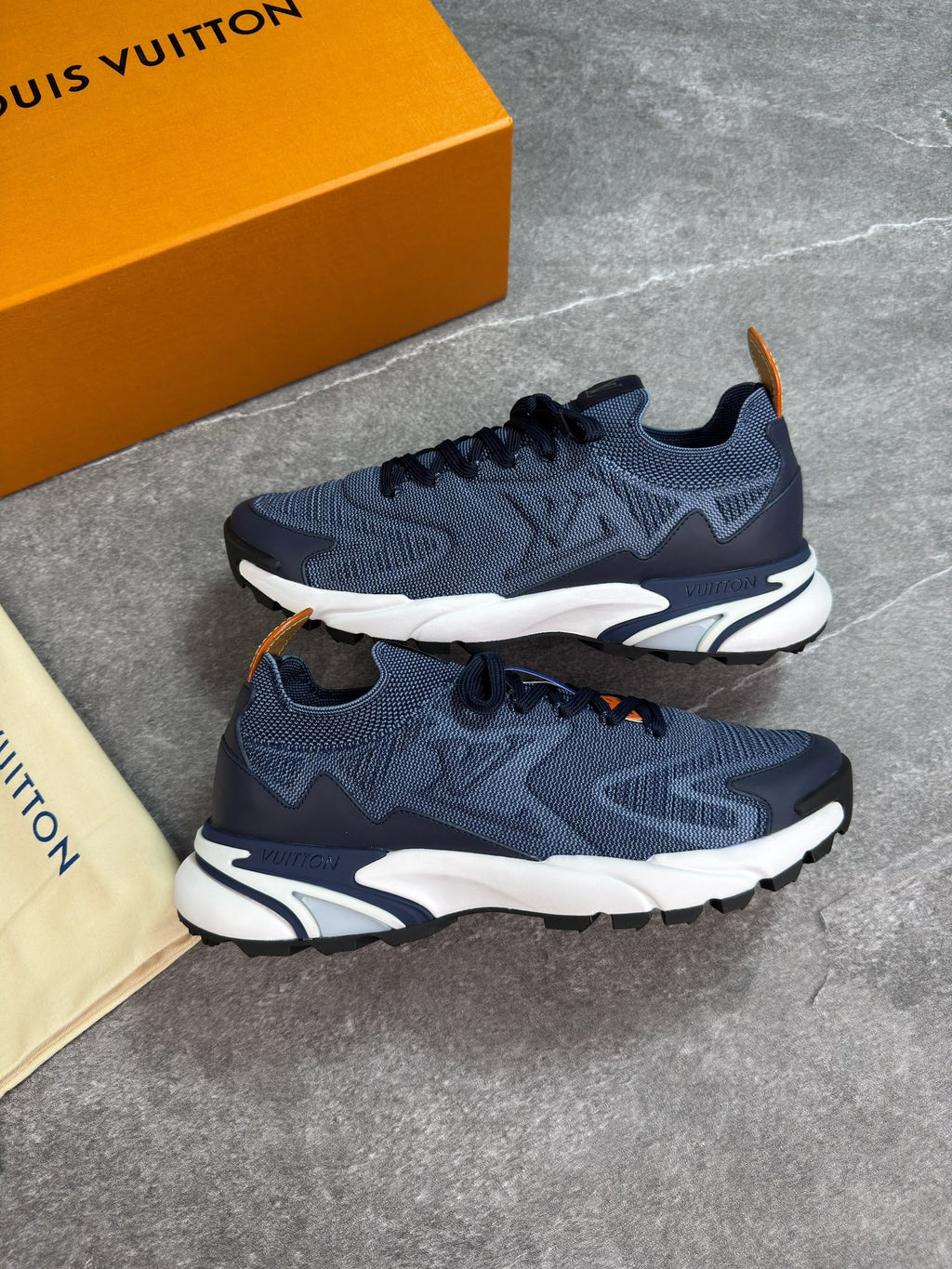 A pair of Louis Vuitton low-top sneakers in navy blue mesh with dark navy overlays, white midsoles, and small orange accents, displayed on a gray stone surface next to an orange Louis Vuitton box and a beige dust bag.