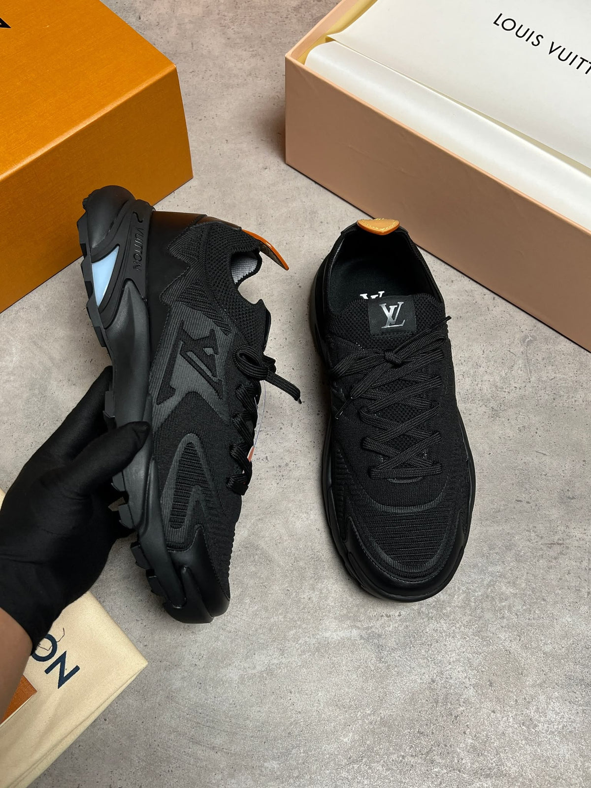 A pair of black Louis Vuitton sneakers shown on a gray floor, with one shoe held by a gloved hand and branded orange and beige boxes visible around them.