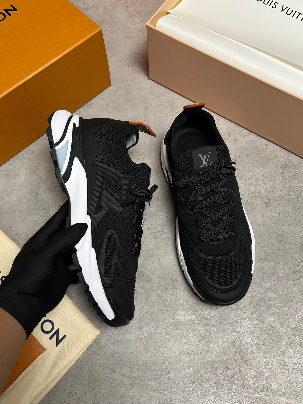 A pair of black Louis Vuitton sneakers with white and orange accents, shown with a black-gloved hand holding one shoe, alongside branded orange and beige shoeboxes on a gray floor.