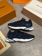 A pair of Louis Vuitton low-top sneakers in navy blue with white and gray midsoles and orange accents, displayed on a gray concrete floor alongside Louis Vuitton boxes and packaging.