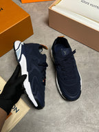 A pair of Louis Vuitton sneakers in navy blue and white, with one shoe being held by a gloved hand and the other placed on a gray floor. Louis Vuitton boxes and packaging are visible in the background.