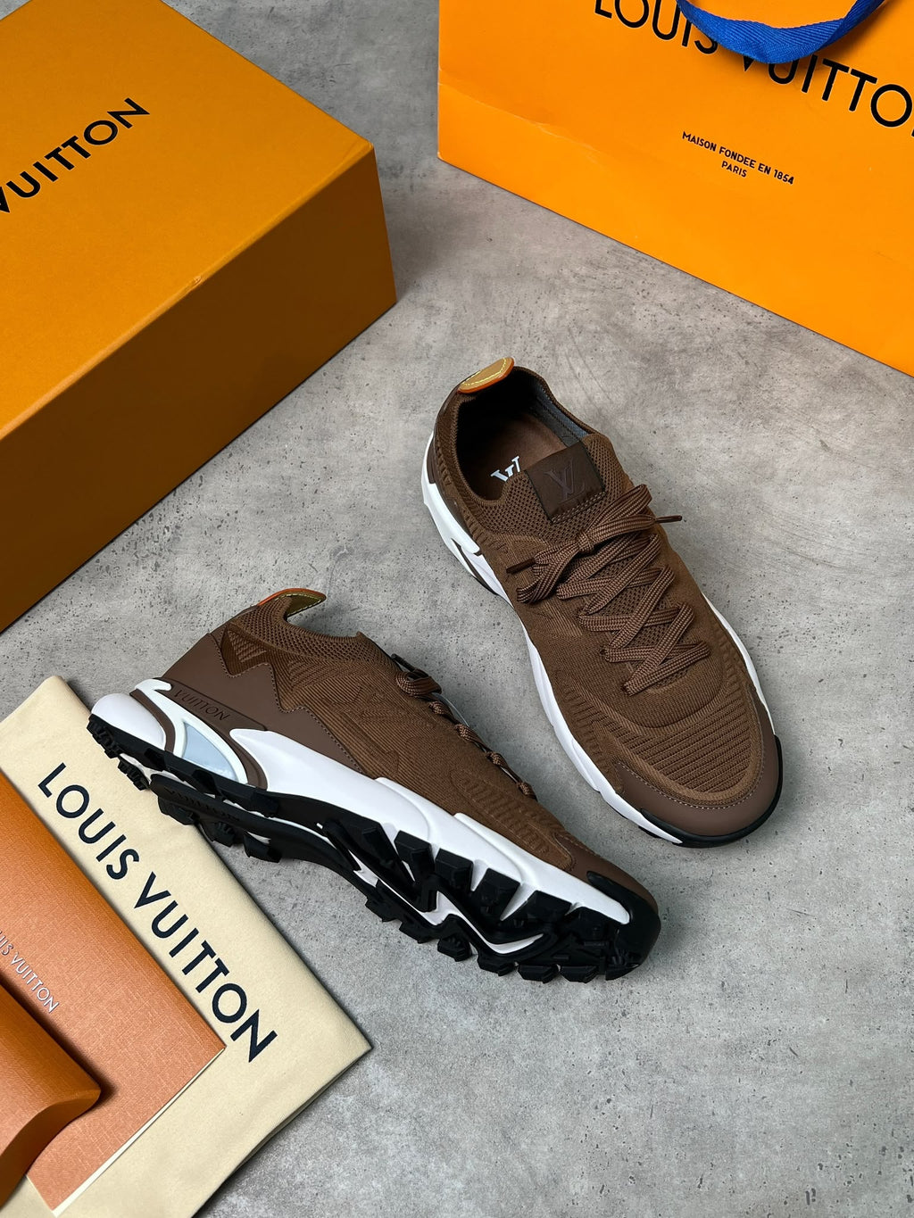 A pair of brown Louis Vuitton sneakers displayed on a gray concrete surface with orange Louis Vuitton boxes and a branded card.