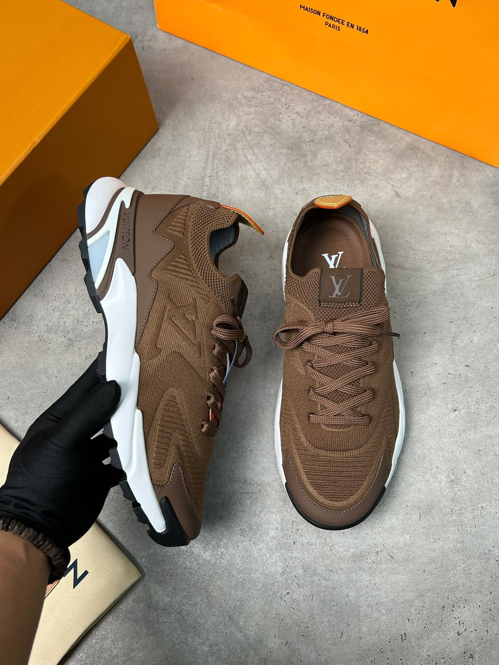 A pair of brown Louis Vuitton sneakers, with one held by a gloved hand and the other placed on a gray concrete floor. Orange Louis Vuitton shoeboxes are visible in the background.