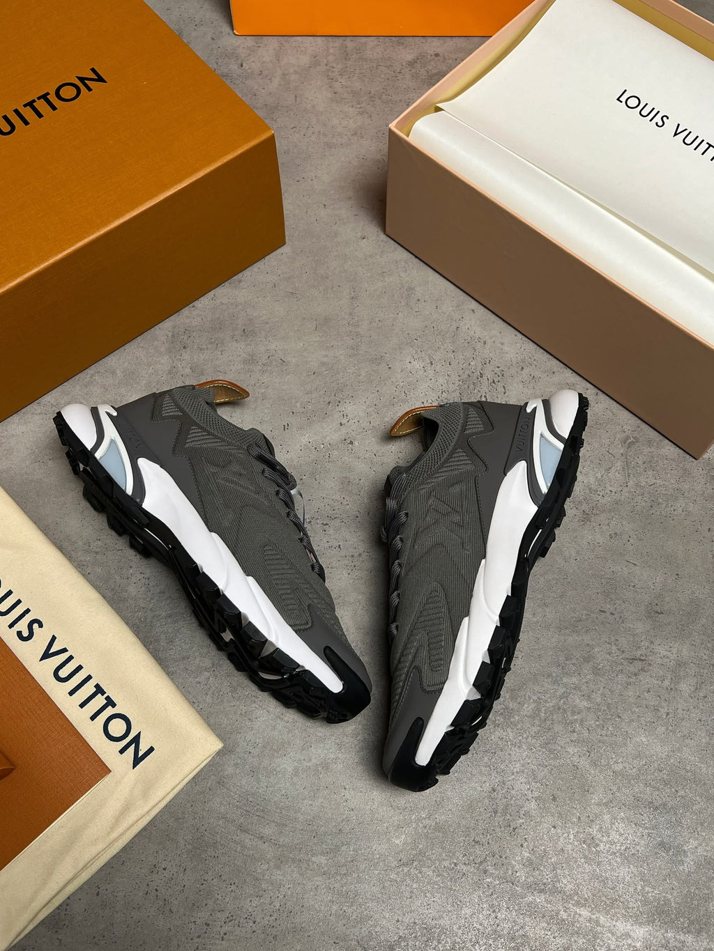 A pair of Louis Vuitton low-top sneakers in dark gray and white, displayed on a gray concrete floor alongside their signature orange and beige Louis Vuitton boxes and dust bags.