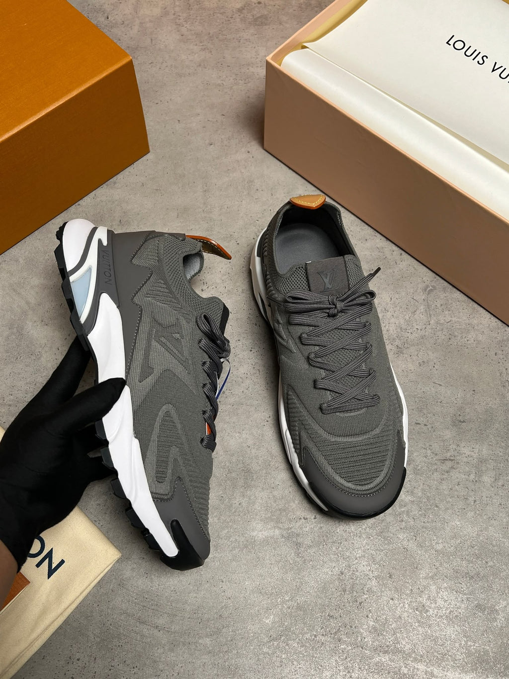 A pair of dark gray and white Louis Vuitton sneakers shown on a gray concrete floor, with one shoe held by a black-gloved hand and branded orange and cream shoeboxes visible in the background.