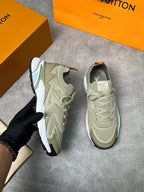 A pair of Louis Vuitton sneakers in a light khaki-beige colorway, with one shoe held by a hand wearing a black glove and the other resting on a gray concrete surface. Orange Louis Vuitton boxes are visible in the background.