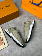 A pair of Louis Vuitton low-top sneakers in beige and white with black soles, displayed on a gray concrete surface alongside an orange shoebox, an orange Louis Vuitton shopping bag, and a small Louis Vuitton card.