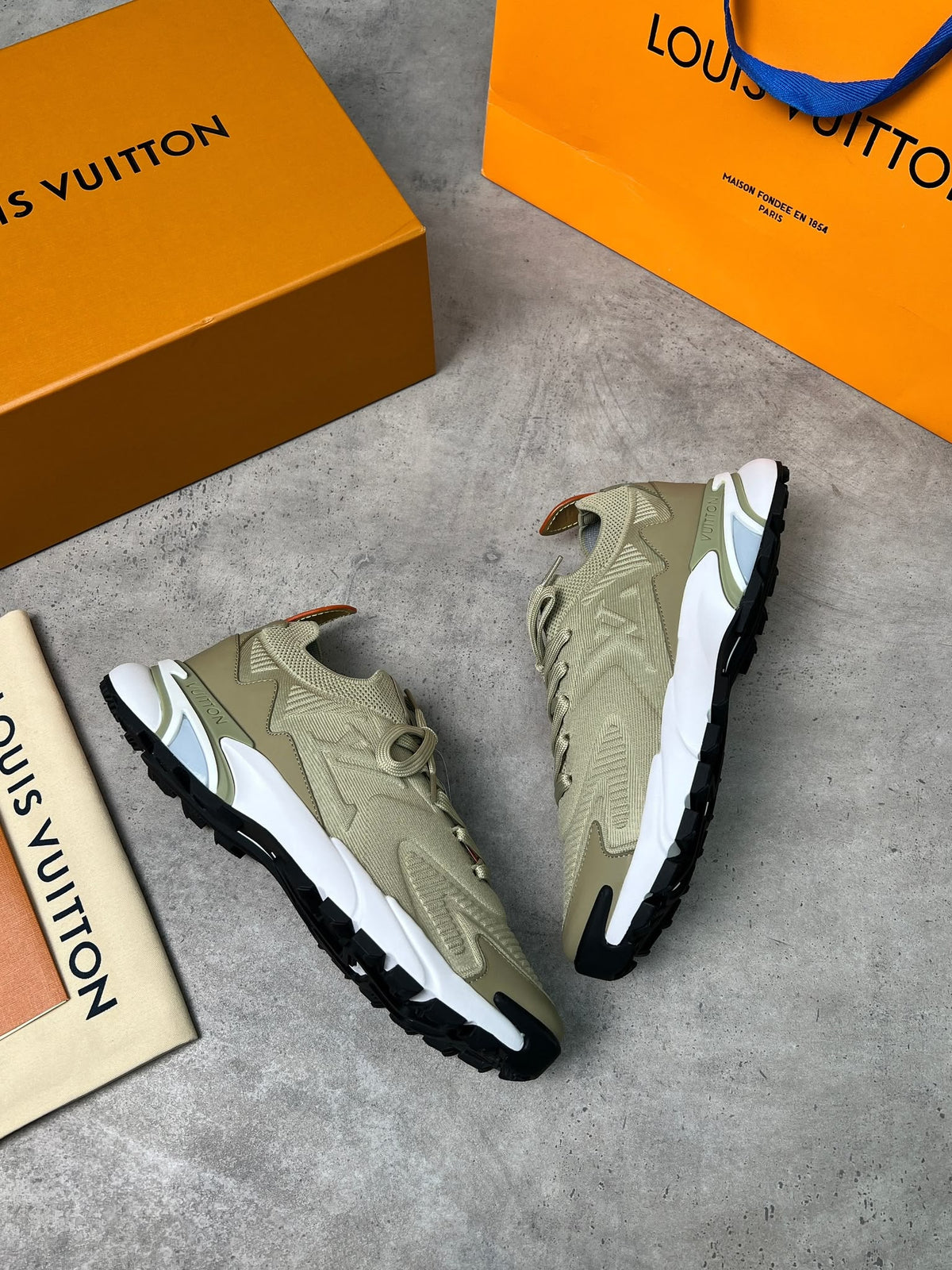 A pair of Louis Vuitton low-top sneakers in beige and white with black soles, displayed on a gray concrete surface alongside an orange shoebox, an orange Louis Vuitton shopping bag, and a small Louis Vuitton card.