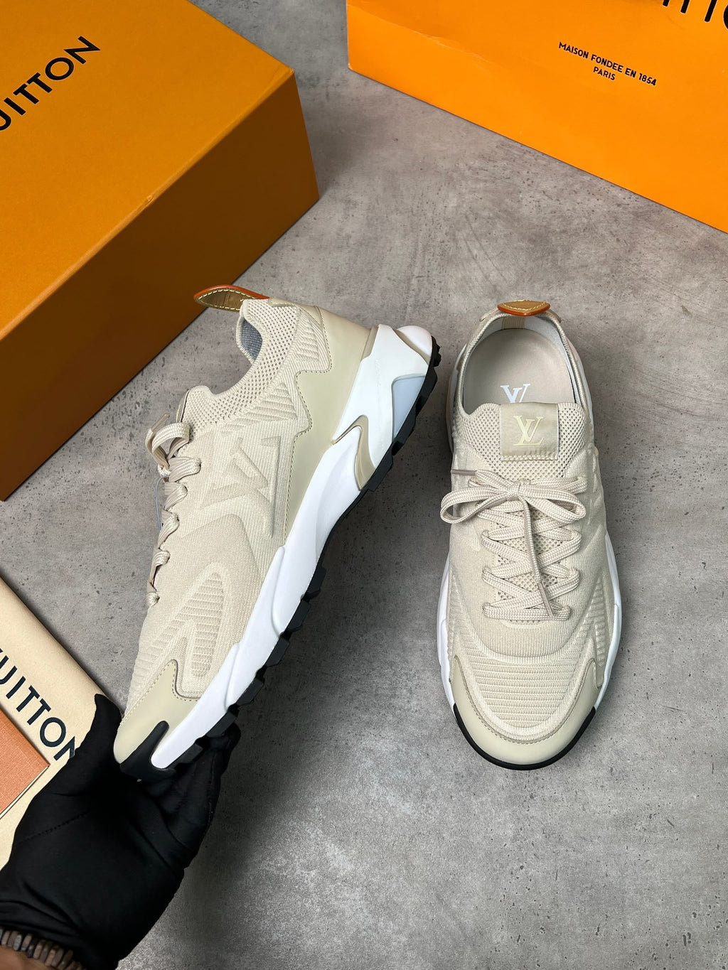 A pair of beige and white Louis Vuitton sneakers shown on a gray concrete floor, with orange Louis Vuitton boxes in the background and a black-gloved hand holding one shoe.