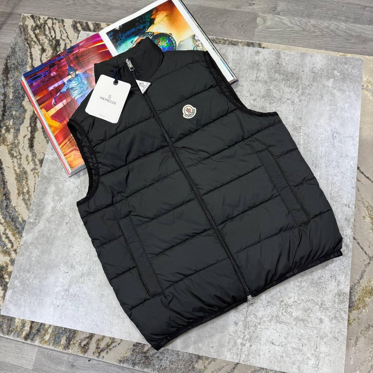 A black Moncler quilted puffer vest laid flat on a gray stone surface, with a white Moncler hangtag attached near the collar and a colorful magazine underneath at the upper left.