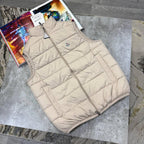 A sleeveless beige puffer vest with a front zipper and a small circular chest logo, laid flat on a gray stone surface next to a colorful magazine.