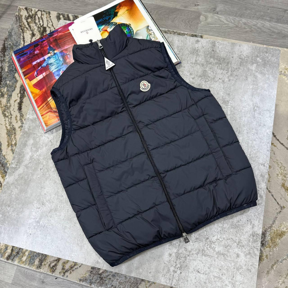 A black puffer vest laid flat on a gray stone slab with colorful magazines underneath. The vest has horizontal quilting, a full front zipper, and a small circular chest logo.