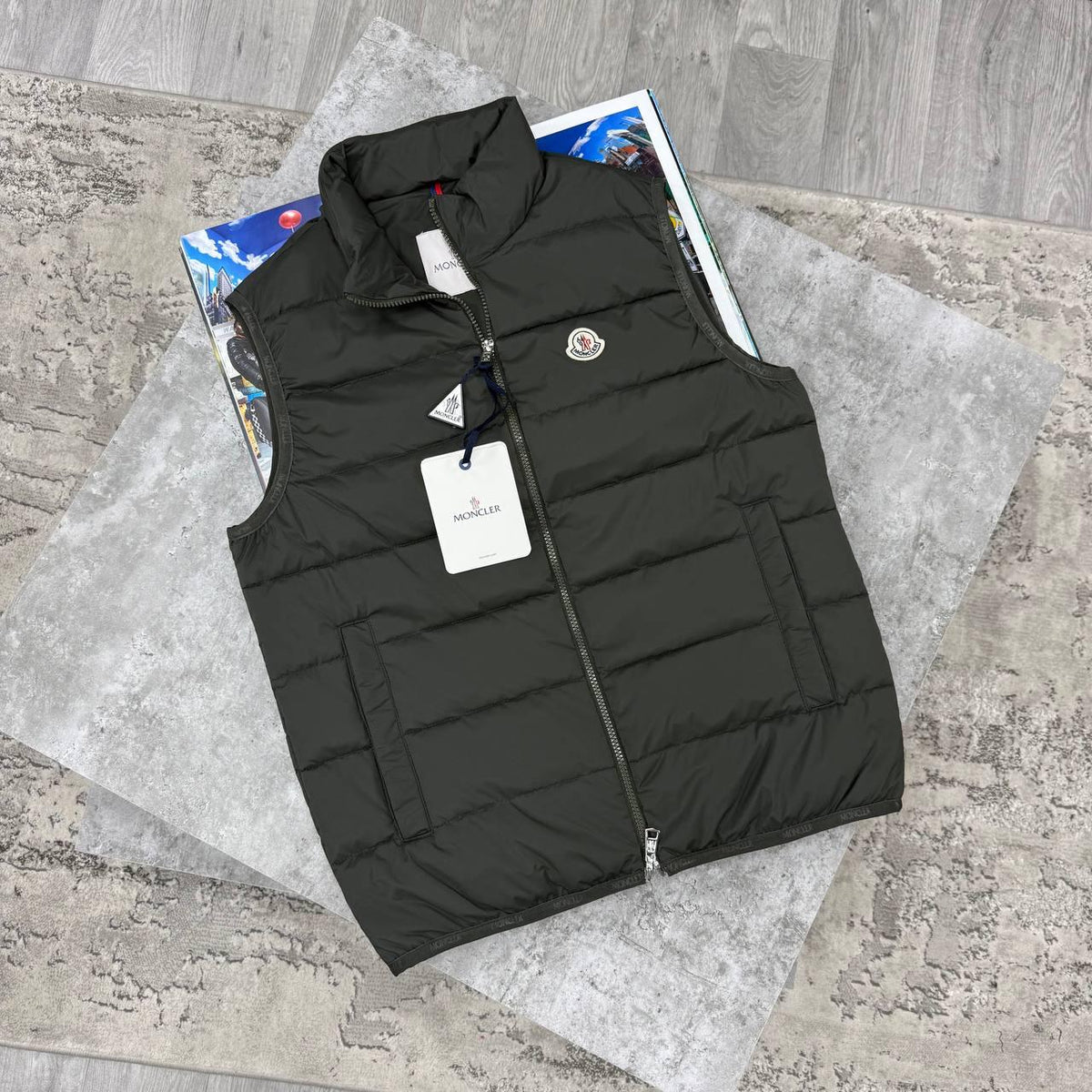 A black Moncler quilted puffer vest laid flat on a gray textured surface, with the brand's circular logo patch on the left chest and a hangtag still attached.