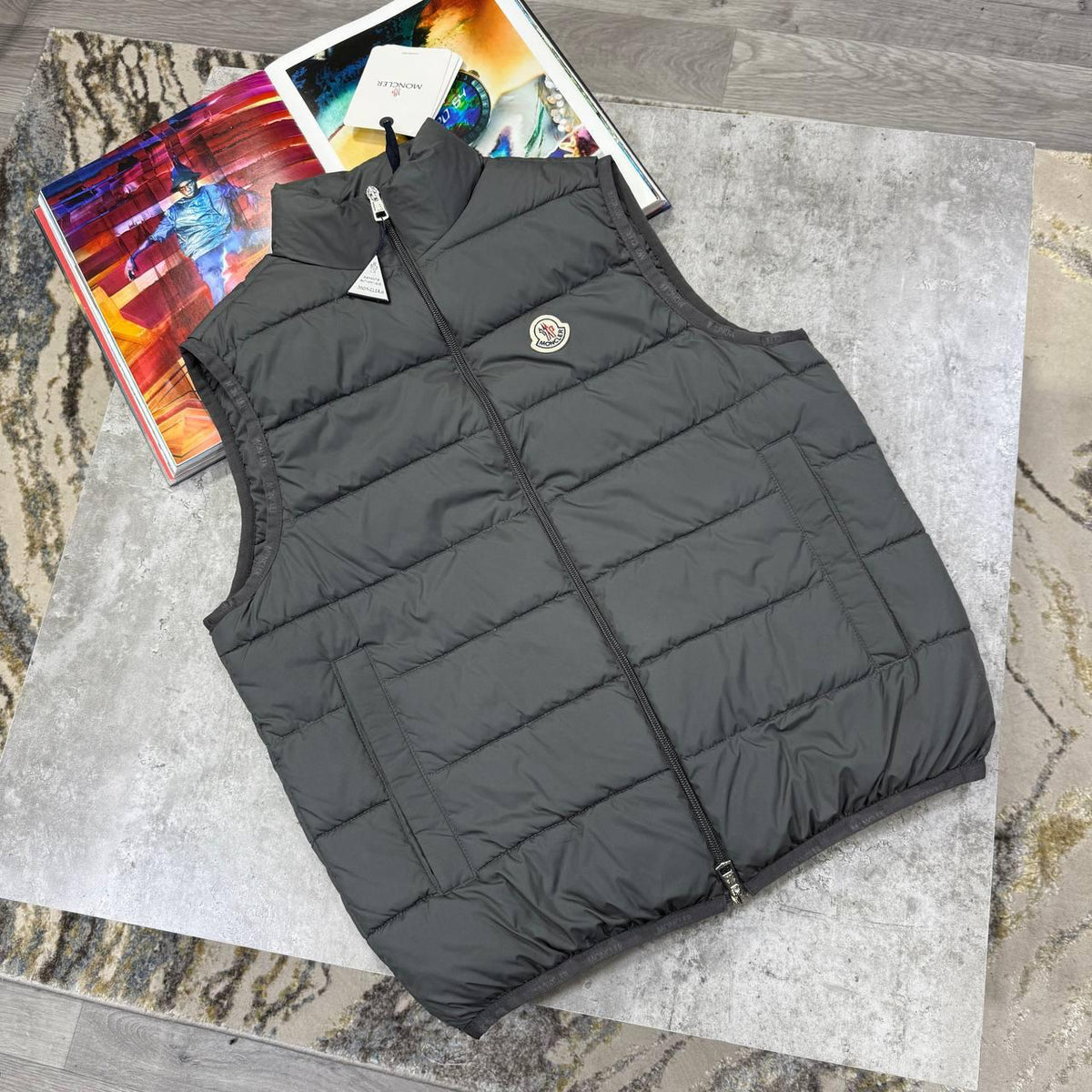 A dark gray sleeveless puffer vest with a full zipper and a small circular chest logo, laid flat on a gray stone surface with books underneath.