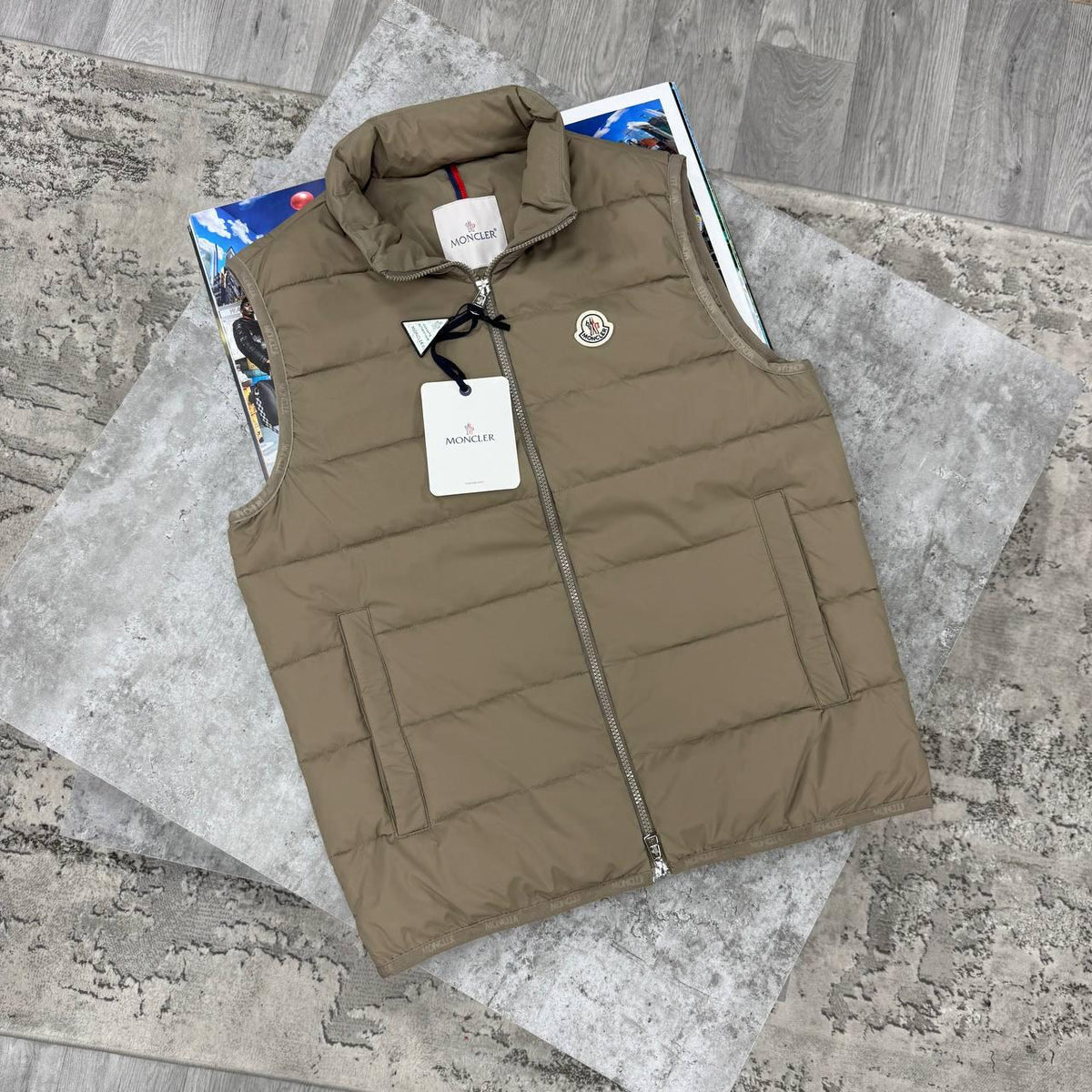 A tan Moncler quilted puffer vest laid flat on a gray stone-patterned mat, with a visible Moncler hangtag and circular chest logo.