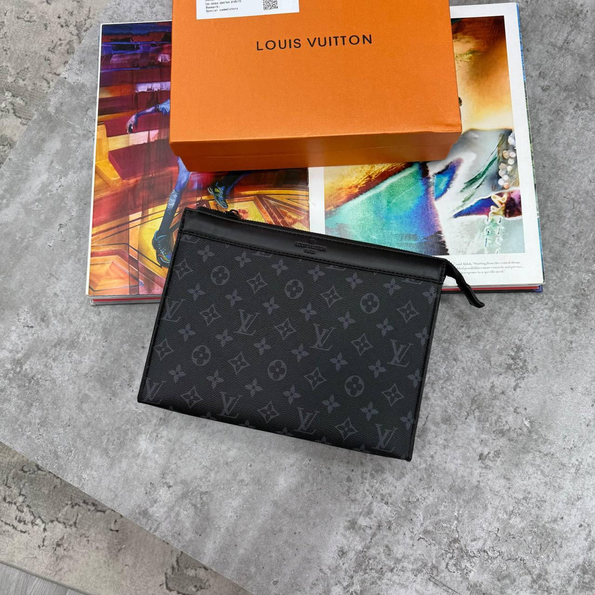 A Louis Vuitton black monogram pouch or small bag placed on top of an orange Louis Vuitton box and colorful printed cards, set on a gray concrete surface.