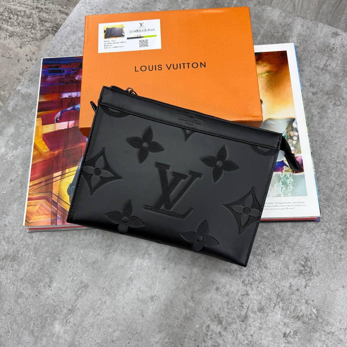 A black Louis Vuitton monogram pouch bag resting on top of an orange Louis Vuitton box and colorful magazines on a gray stone surface.