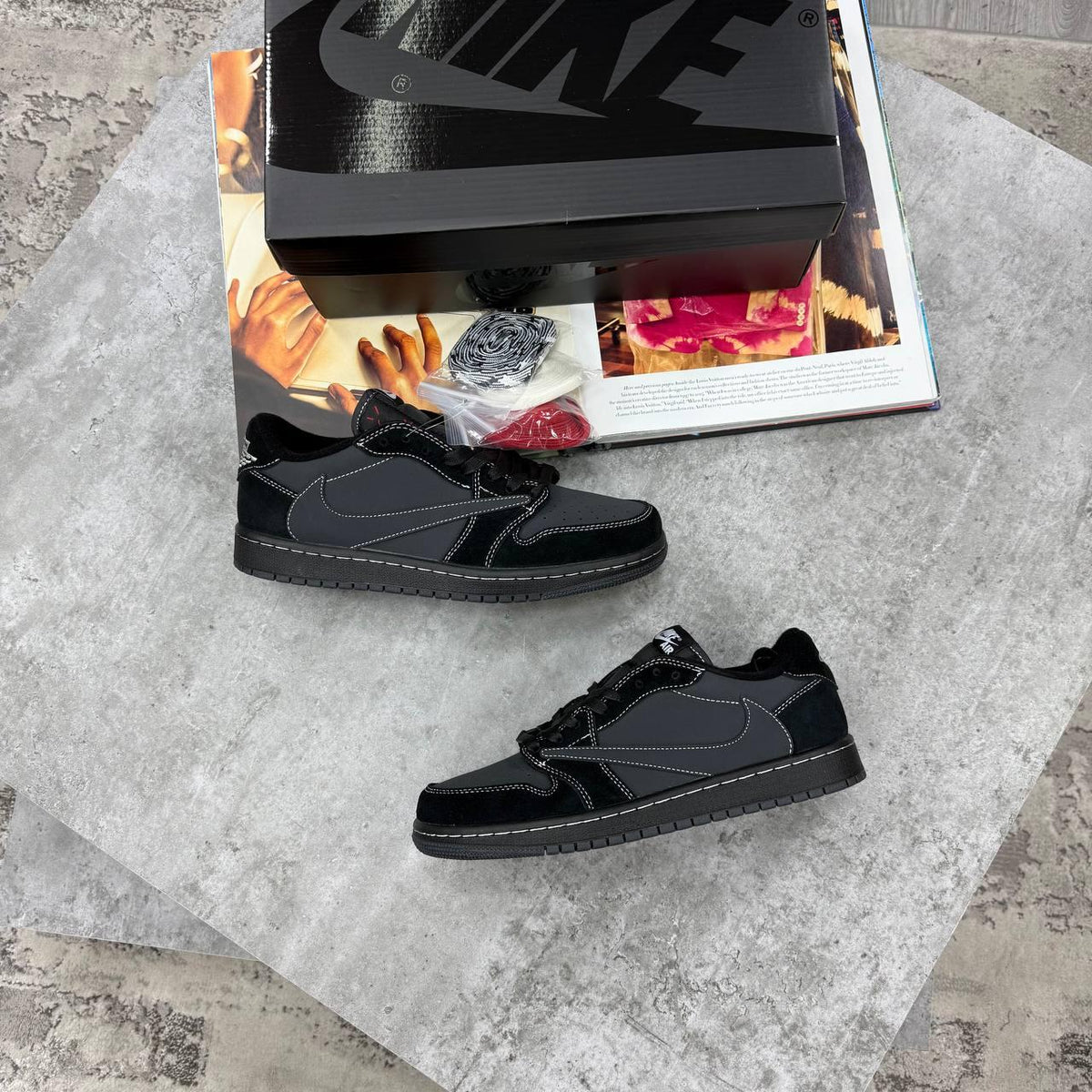 A pair of black Nike low-top sneakers displayed on a gray stone surface with an open magazine and a black Nike shoebox behind them.