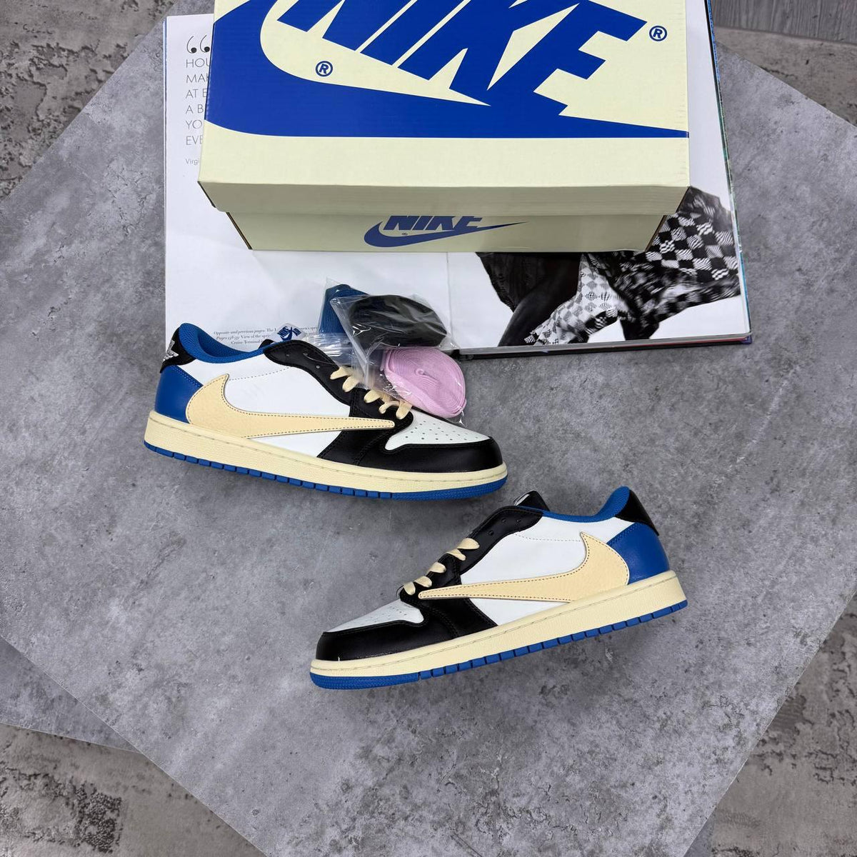 A pair of Nike Dunk Low sneakers in a black, cream, white, and blue colorway, displayed on a gray concrete surface with their original box and a magazine in the background.