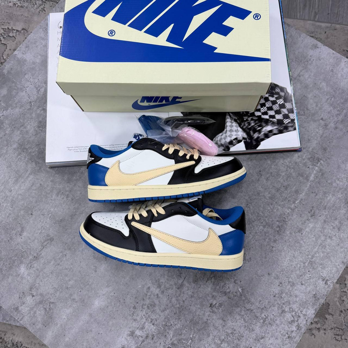 A pair of Nike low-top sneakers in a black, cream, and blue colorway, displayed on a gray stone surface with their branded shoebox and a magazine underneath.